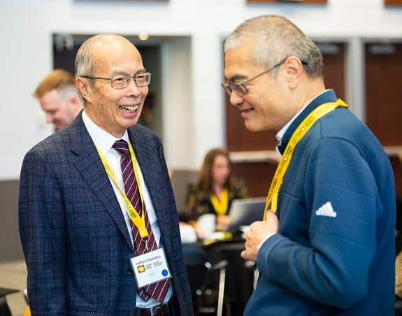 Two individuals talk with each other at an indoor event, wearing name tags and standing in front of other seated attendees.