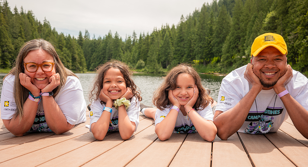 wo adults and two children lie side by side on a wooden dock overlooking a calm lake surrounded by forested hills.