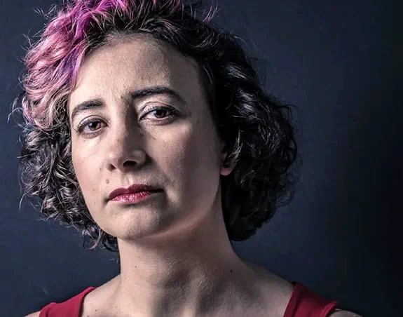 A woman in her 40s with a serious expression makes eye contact.