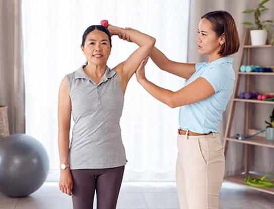 A woman has her arm raised and bent at the elbow to hold a dumbbell behind her head, guided by a physiotherapist