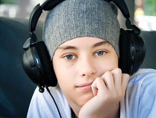 A male teenager is wearing headphones while sitting with his hand on his chin. 