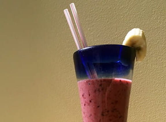 A fruit smoothie with two straws and a slice of banana on the rim of the glass