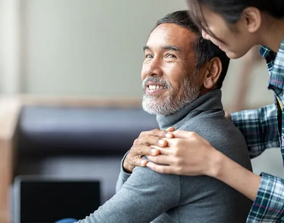 A woman standing behind a man with her hand on his shoulder. He is looking back at her, his hand on top of hers and smiling.