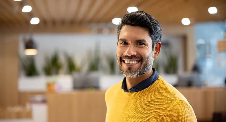 Un homme debout dans un bureau et souriant en regardant en direction de l’appareil photo.