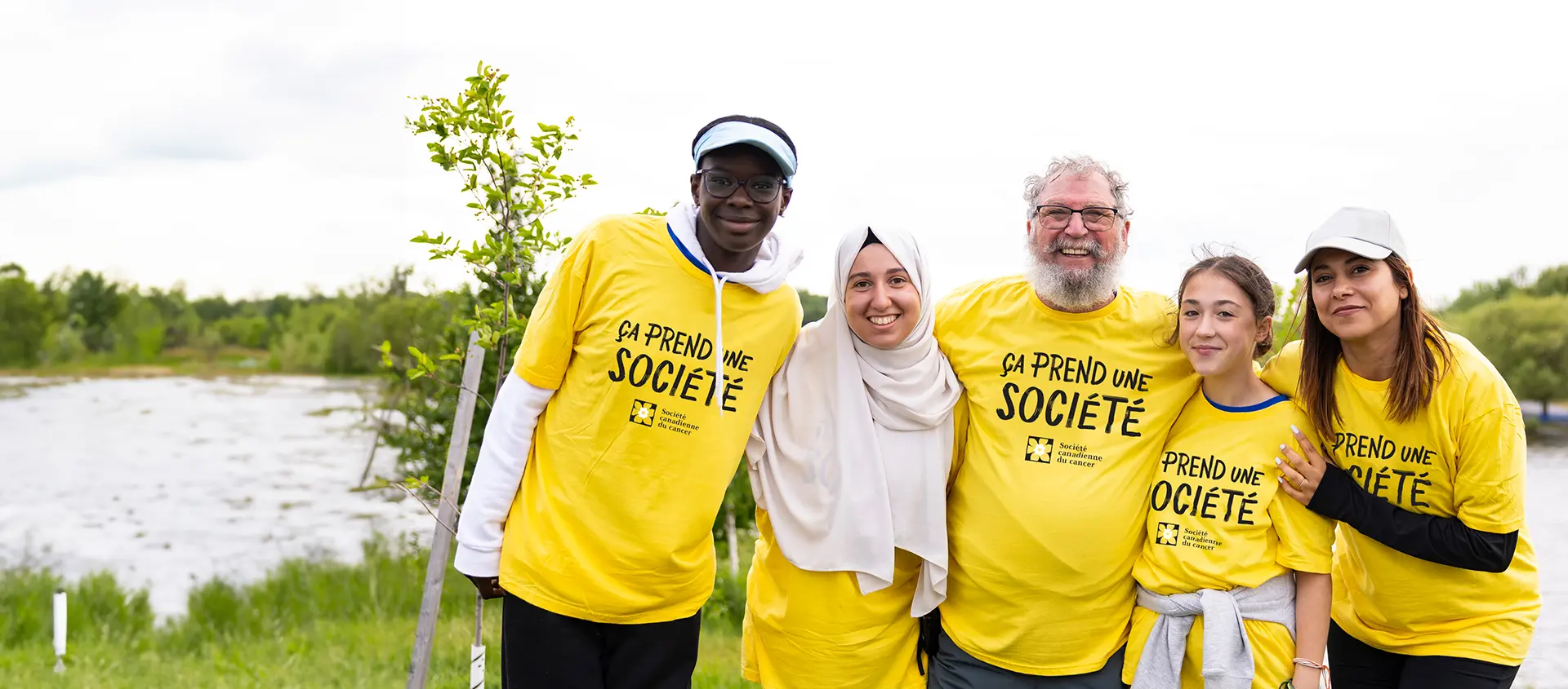 Un groupe de personnes portant des T-shirts « Il faut une société » se tient devant une zone boisée et sourit.