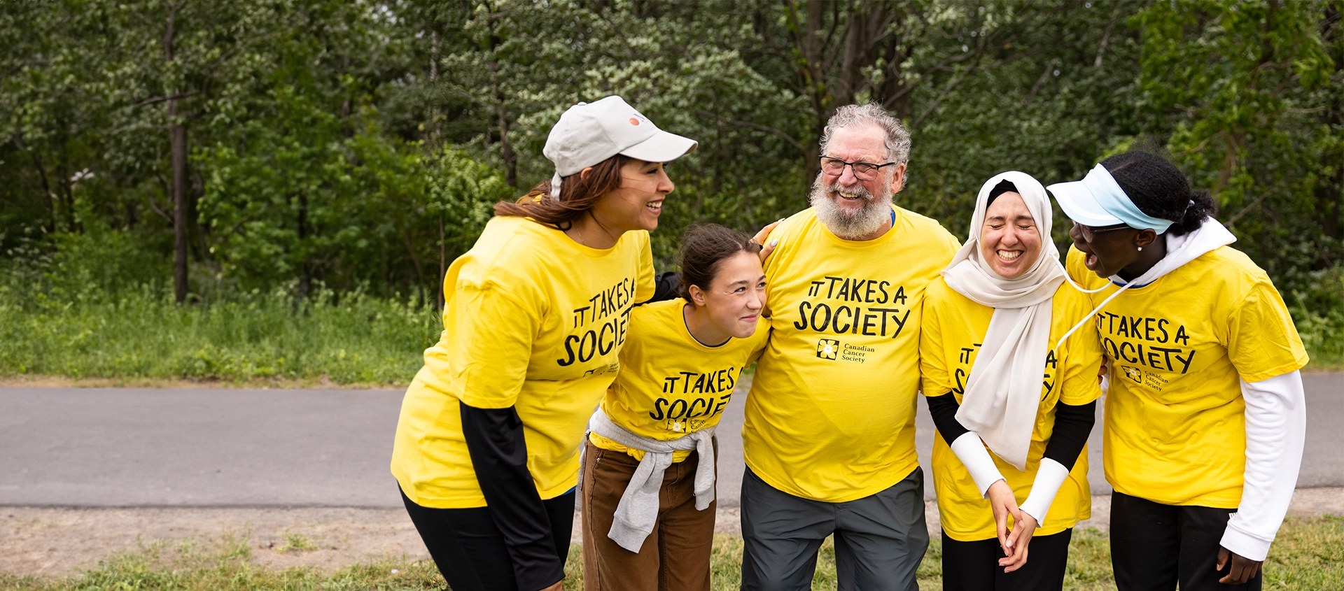 A group of people wearing ‘It takes a society” T-shirts are standing in front of a wooded area. They are laughing and smiling.