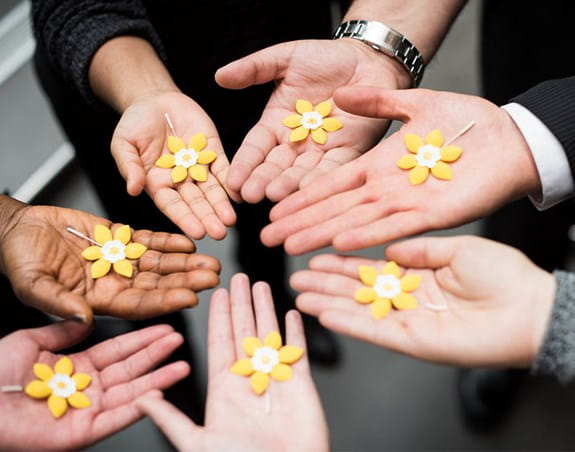Seven people with their hands in a circle with their palms up, each holding a daffodil pin.