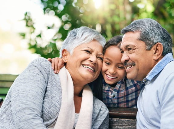 Two grandparents sit on a park bench hugging their granddaughter