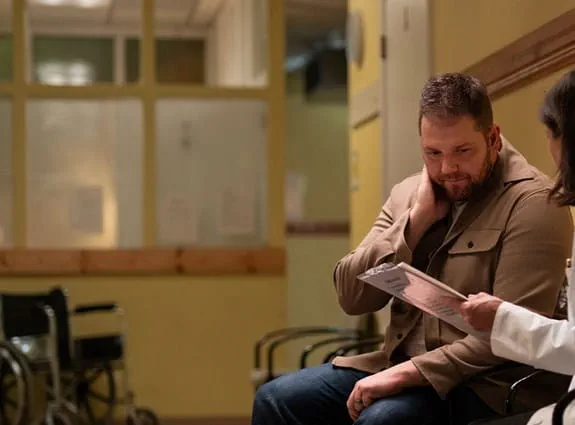 A doctor is holding a medical chart and talking to a person as they sit in a hospital hallway.