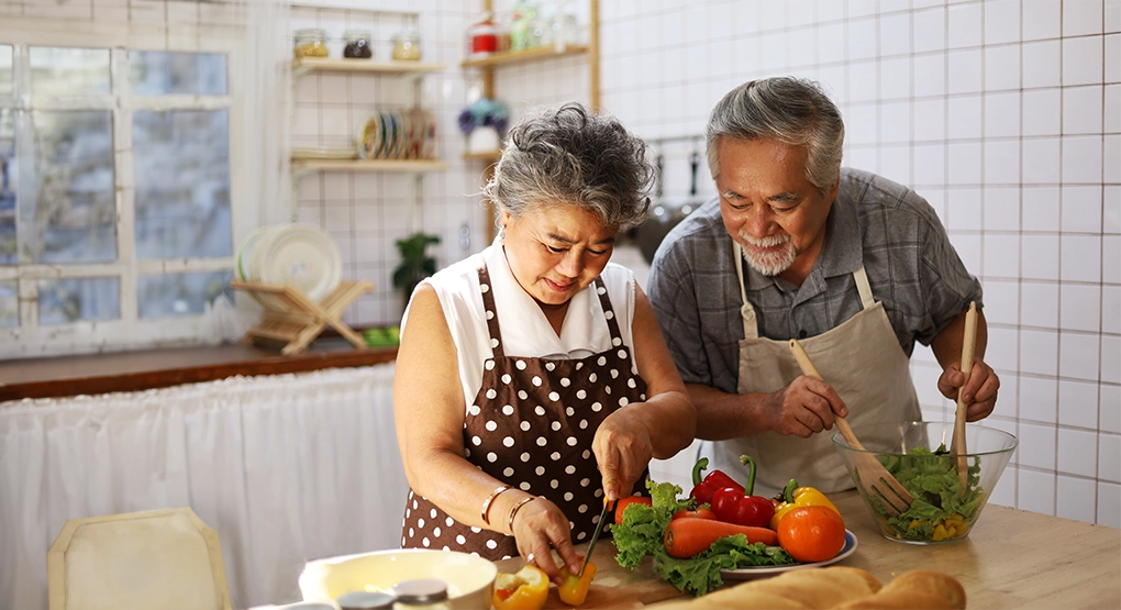 An elderly couple in a kitchen preparing a salad with fresh vegetables.