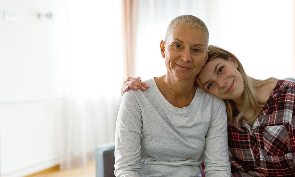 A woman sits on a couch with her arms around an older woman. They are both smiling.