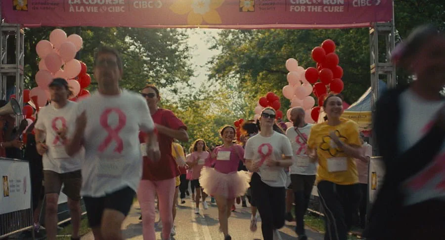 Participants running in a charity race under a banner that reads 'CIBC Run for the Cure' and 'La Course à la vie CIBC,' with festive balloons and trees in the background.