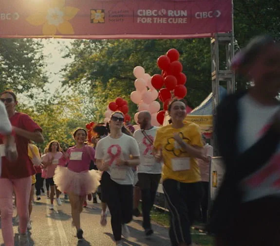 Participants running in a charity race under a banner that reads 'CIBC Run for the Cure' and 'La Course à la vie CIBC,' with festive balloons and trees in the background.