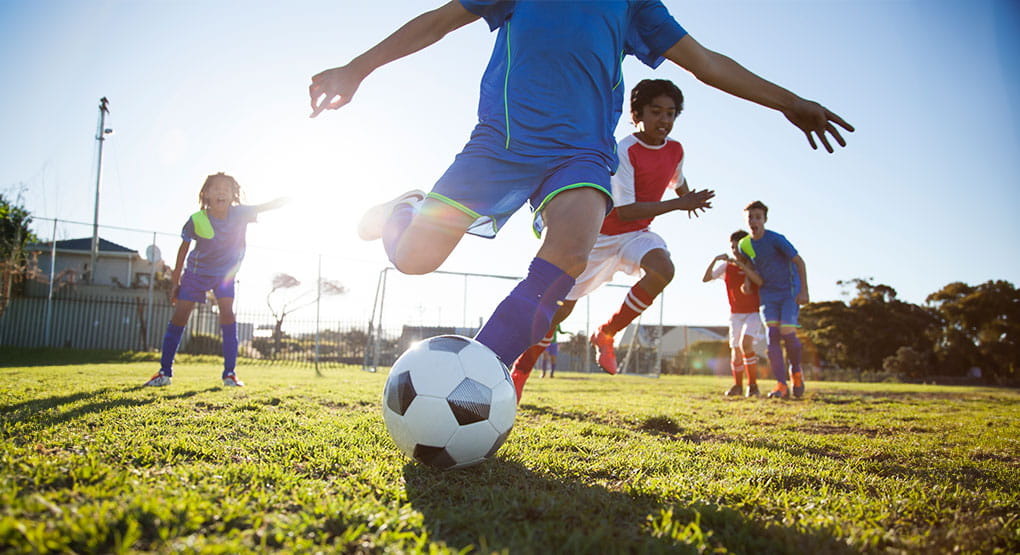 Several people playing soccer on a grassy field, with one person kicking a soccer ball in the foreground while others run nearby during a Soccer for Cancer fundraiser event.