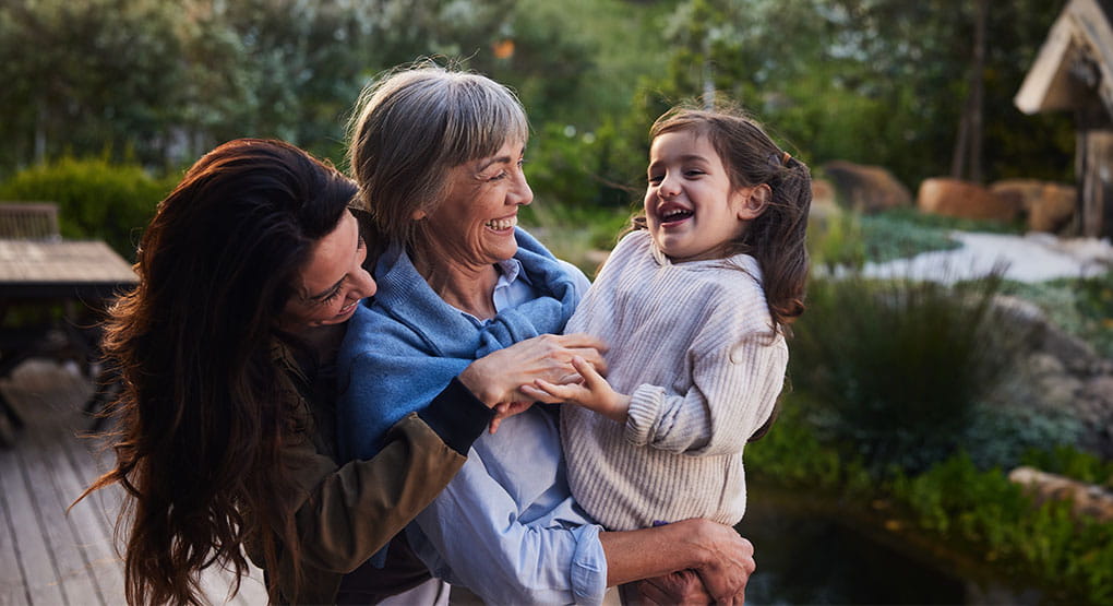 A mother and daughter standing close together while holding a young child in a garden setting.