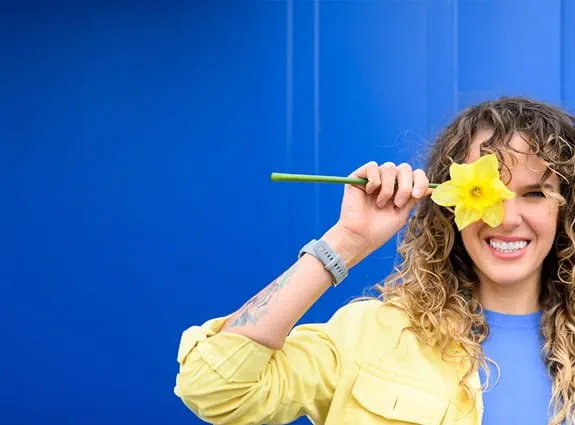 A woman with a bright smile holds a daffodil flower over her right eye, looking directly at the camera.