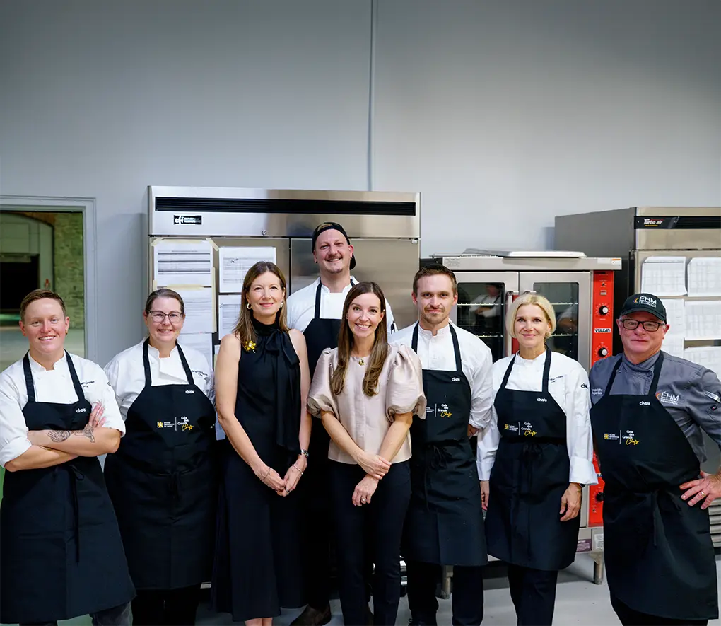 A group of eight people stands in a professional kitchen, some wearing chef uniforms and aprons.