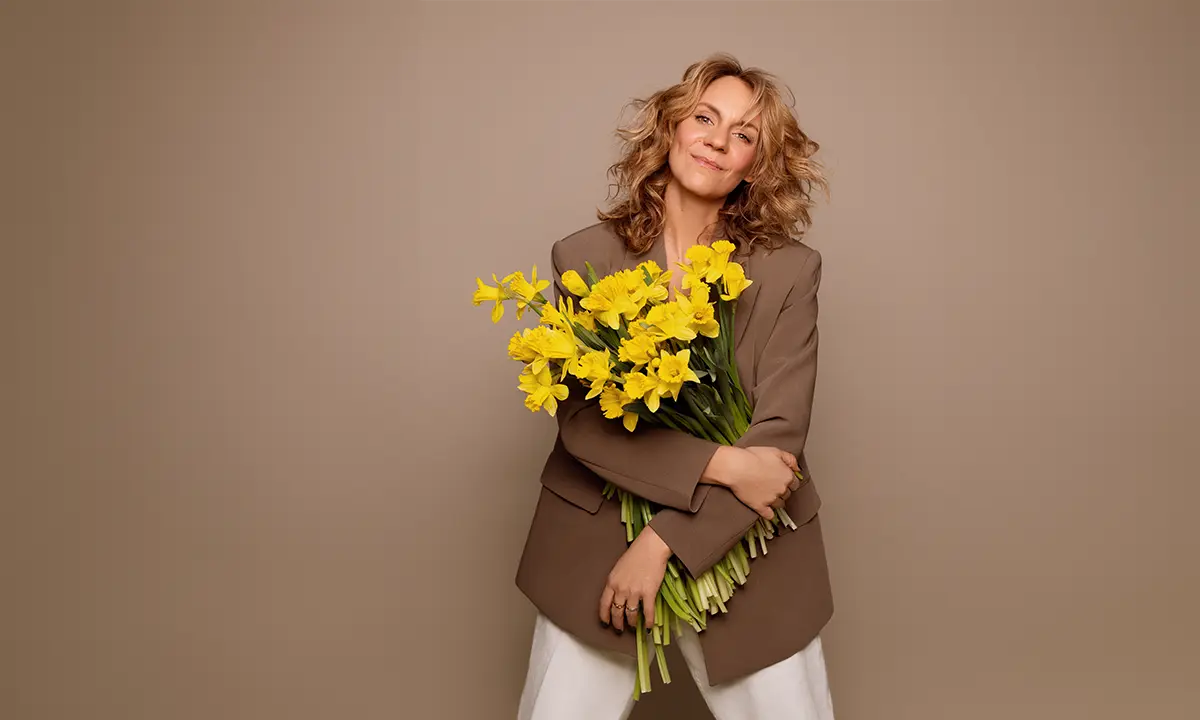Léane Labrèche-Dor standing, holding a bouquet of yellow daffodils, wearing a brown blazer, against a neutral background for CCS’s campaign