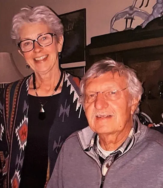 A husband and wife smiling. Both are in casual attire and wear glasses.