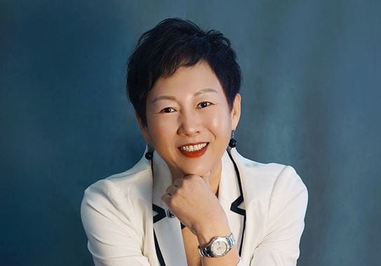 Une femme souriante aux cheveux courts est assise dans un studio de photographie. Elle porte une veste blanche et un jean. 