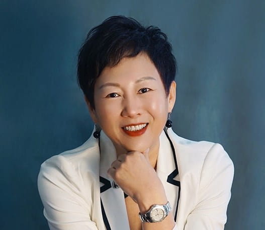 Une femme souriante aux cheveux courts est assise dans un studio de photographie. Elle porte une veste blanche et un jean. 