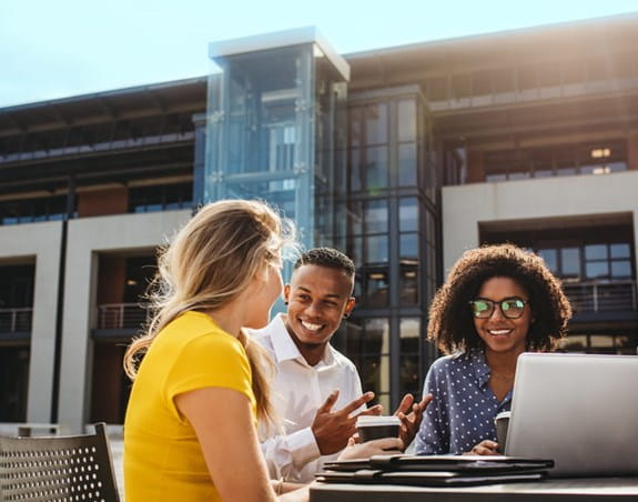 Four people sit outside working at a table, they are smiling as they talk with each other