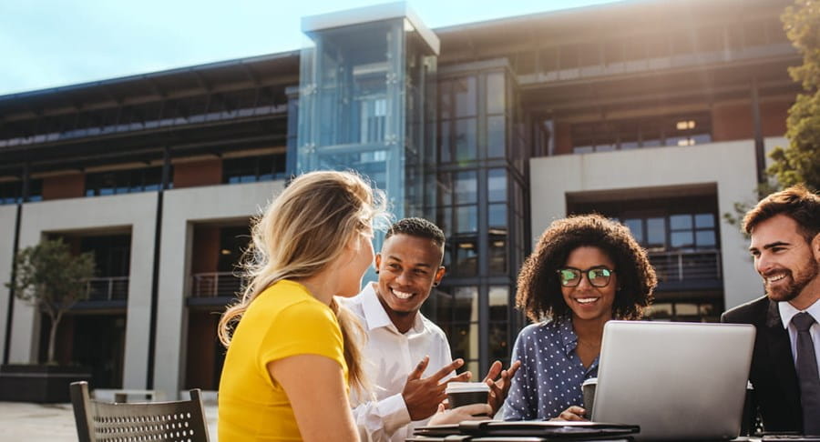 Four people sit outside working at a table, they are smiling as they talk with each other