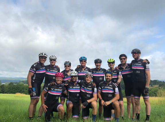 A group of Randstad Ride for Myriam cyclists smiling at the camera while standing in a field.