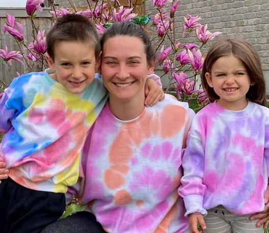 Alexis et ses deux enfants dans leur jardin, portant des chandails tie dye et souriant.   