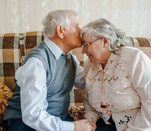 An elderly couple sits on a sofa, leaning towards each other and holding hands while the man is kissing his wife on the forehead.