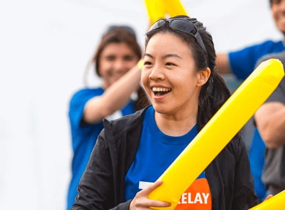 A volunteer is smiling and clapping a pair of inflatable thundersticks, others clap in the background.