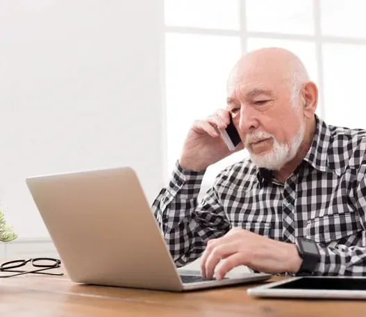 Older man typing on a laptop with a smartphone held to his ear