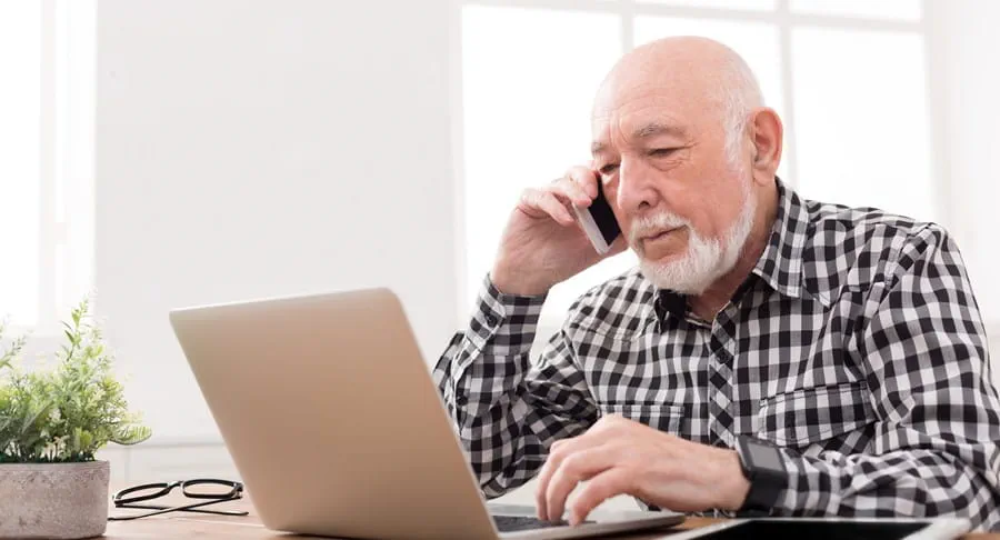 Older man typing on a laptop with a smartphone held to his ear