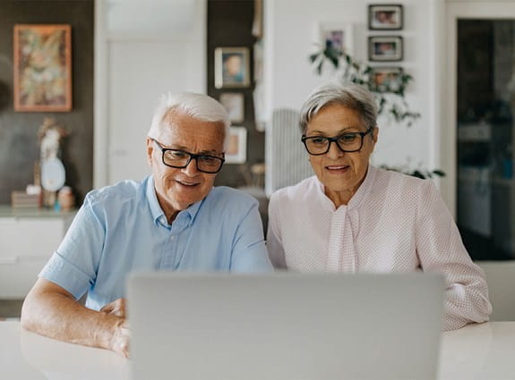 Two older adults sit at a table looking at a laptop computer.