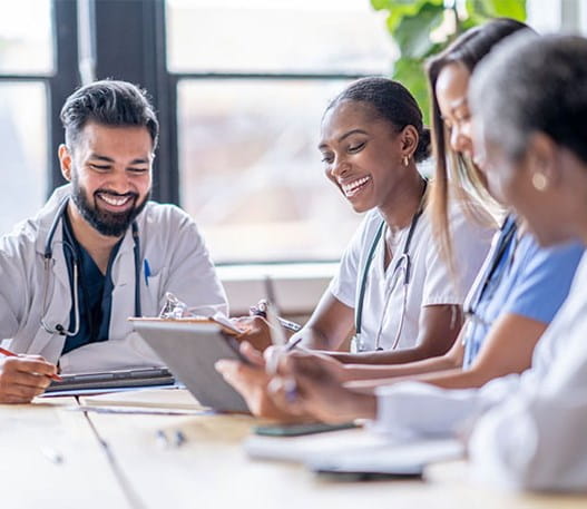 Four healthcare providers sit around a table talking and taking notes while looking at a tablet.