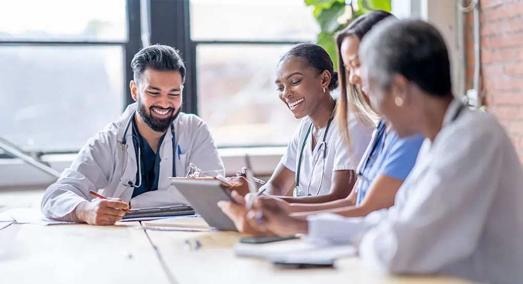 Four healthcare providers sit around a table talking and taking notes while looking at a tablet.