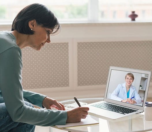 A person takes notes during a video call with their healthcare provider. Two pill bottles are on the table next to them.