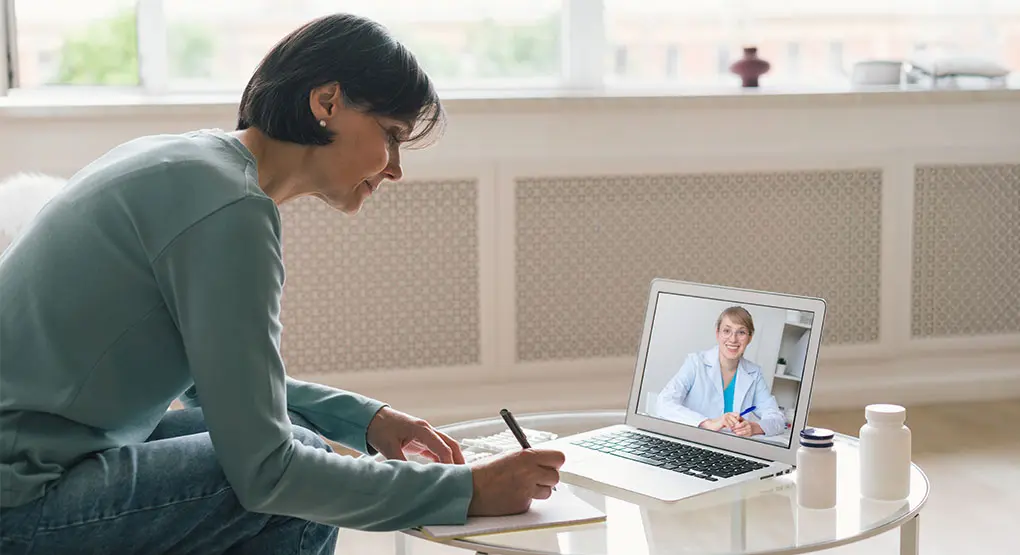 A person takes notes during a video call with their healthcare provider. Two pill bottles are on the table next to them.