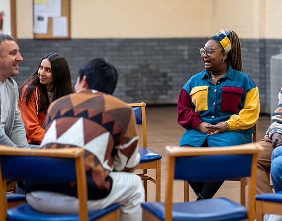 A diverse group of people sit in a circle in a community centre, engaged in a pleasant discussion.