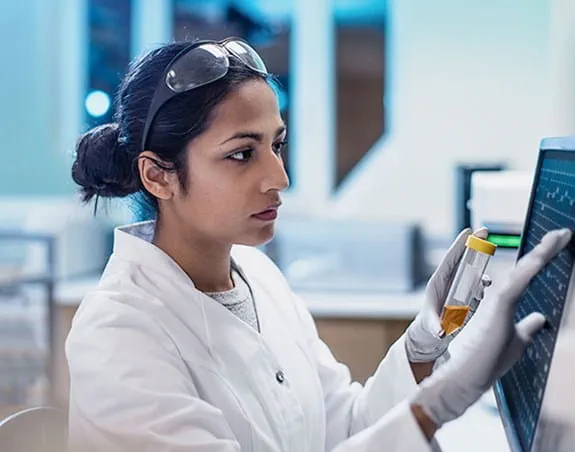 A female cancer researcher typing into a touch screen and holding a vial in a lab.