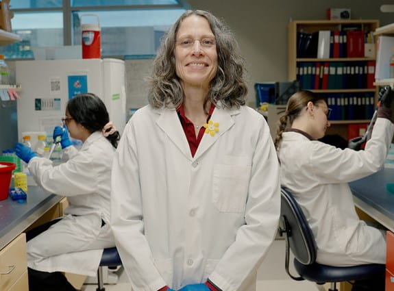 A person in a lab coat smiles in a laboratory while others work in the background.