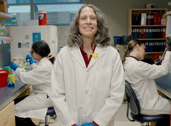 Une personne portant un sarrau sourit dans un laboratoire pendant que d’autres travaillent en arrière-plan.