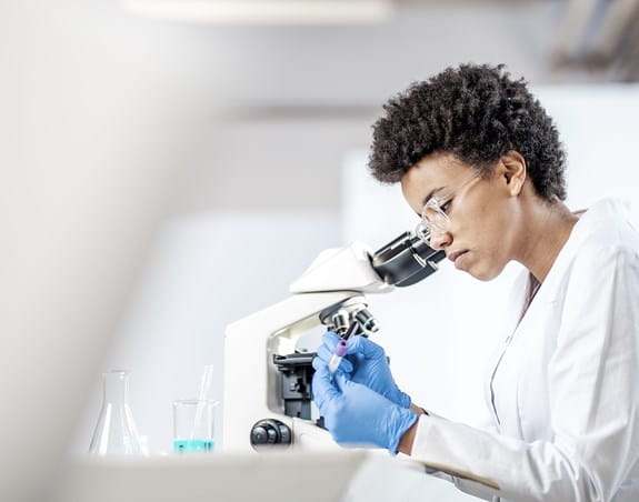 A female scientist is writing on a test tube while sitting behind a microscope