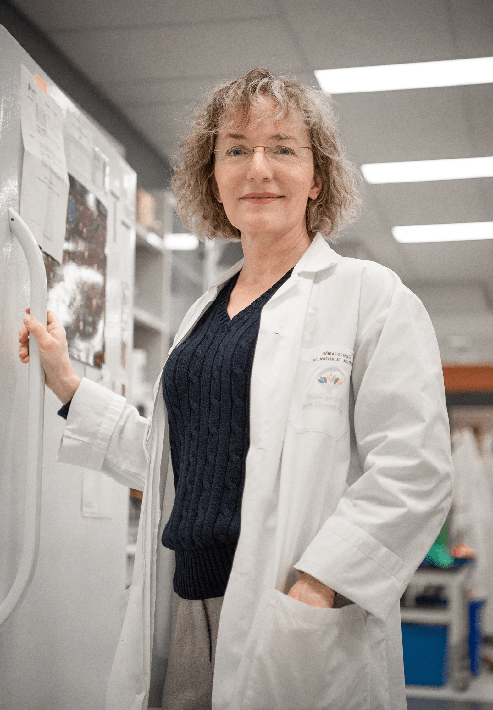 La Dre Nathalie Johnson, une femme à la peau claire, aux cheveux blonds et vêtue d’un sarrau, est debout et tient la poignée du réfrigérateur du laboratoire  