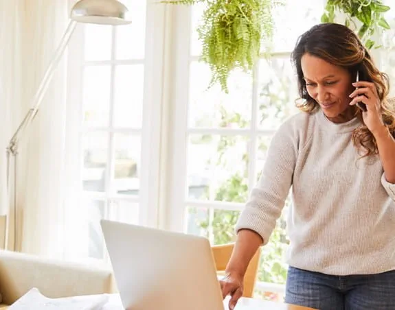 A woman is speaking on her phone while looking at a laptop in her living room