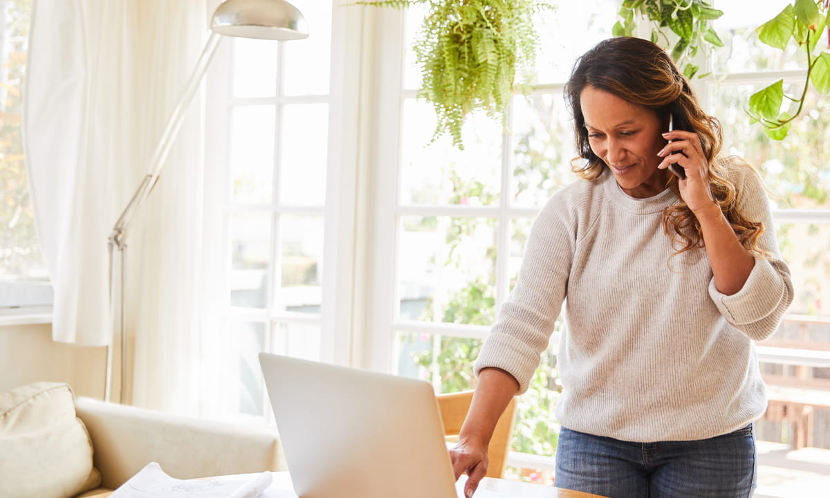 Une femme dans son salon, parlant au téléphone en regardant l’écran de son ordinateur portable.