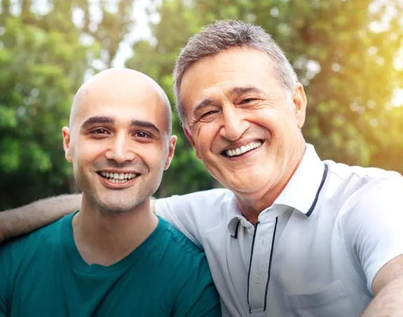 young man with a beard smiling looking over at an older man, sitting on a couch