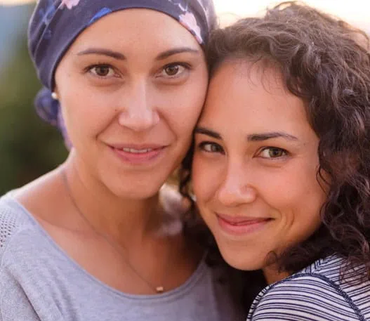 A smiling woman with cancer wearing a scarf on her head with another woman.