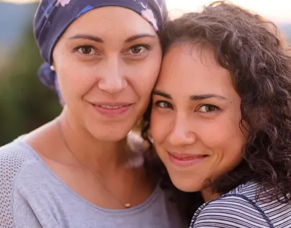 A smiling woman with cancer wearing a scarf on her head with another woman.