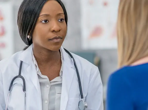 A female doctor wearing a lab coat and stethoscope addressing a patient.